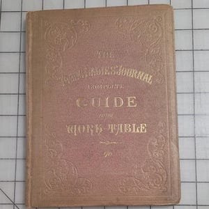 May include: A vintage book titled "The Young Ladies' Journal Complete Guide to the Work Table." The cover is a faded pink with gold lettering and decorative corner designs. The book is rectangular and appears well-worn.