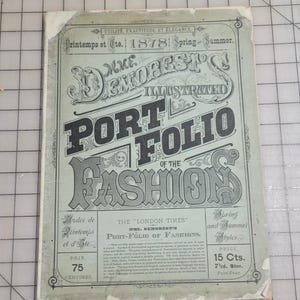 May include: An antique magazine cover titled "Mme. Demorest's Illustrated Port Folio of the Fashion" from 1878. The cover is light green with black text and decorative flourishes. It features text in French and English, advertising spring and summer styles.