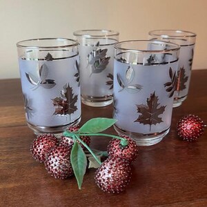 May include: Set of four vintage drinking glasses with a frosted finish and silver leaf design. Each glass has a silver rim. Decorative red and silver cherries with green leaves are in the foreground.