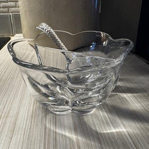 May include: A clear glass serving bowl with a decorative floral design and a matching serving spoon. The bowl has a scalloped edge and sits on a light-colored surface. The spoon has a textured handle.