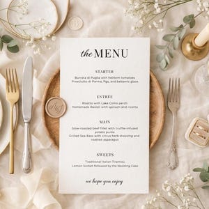 Pode incluir: Menu de casamento elegante com o texto "THE MENU" no topo. O menu inclui entradas, prato principal e sobremesas. O menu é rodeado por pratos, talheres, flores e uma vela acesa.