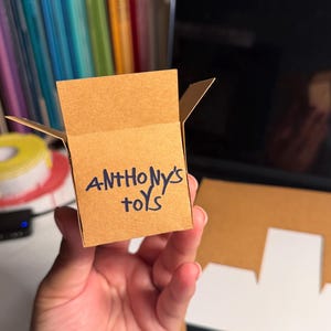 May include: A small, open-top cardboard box made of light brown paper. The front of the box features the handwritten text "ANTHONY'S TOYS" in dark blue ink. The box has four flaps extending from the top edges, creating an open container design.