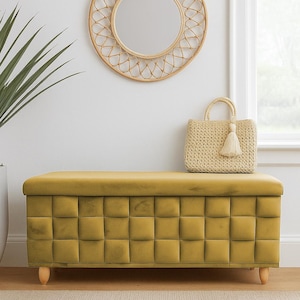 May include: A mustard yellow tufted storage bench with wooden legs. A woven handbag with a tassel sits on top. A round, woven mirror hangs on the wall above the bench. Home decor.