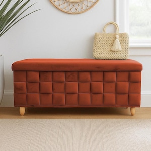 May include: A rust-colored, tufted storage bench with wooden legs. A woven handbag with a tassel sits on top, near a round, woven mirror. The bench is placed against a white wall, with a large plant and window in the background.