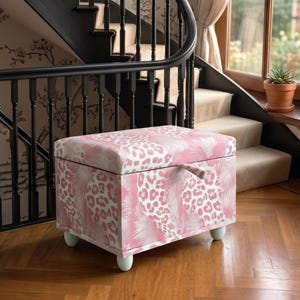 May include: A rectangular storage ottoman featuring a pink and white leopard print pattern. The piece has a hinged lid with a small fabric pull tab and is supported by four rounded, light-colored feet, positioned on a wooden floor.