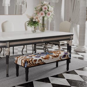 Puede incluir: Un lujoso juego de comedor con una mesa rectangular blanca con ribete negro y patas ornamentadas. Seis sillas color crema rodean la mesa y un banco estampado se encuentra debajo. Un arreglo floral y un espejo completan la elegante escena.