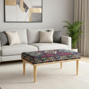 Könnte beinhalten: Ein Wohnzimmer mit einem hellgrauen Sofa, einer gemusterten Ottomane und einem neutralen Teppich. Ein abstraktes Gemälde hängt über dem Sofa, und eine Topfpflanze sorgt für Grün. Der Raum ist hell und natürlich beleuchtet.