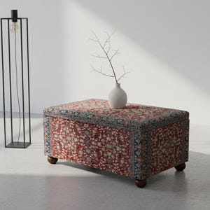 May include: A rectangular ottoman with a floral patterned fabric in red, cream, and blue tones. It has dark wooden legs and a small white vase with a branch on top. A black metal floor lamp with a single exposed bulb stands nearby.