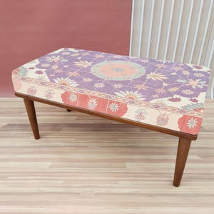Peut inclure: Un ottoman rectangulaire avec un motif floral dans les tons de violet, rose et crème. Les pieds en bois sont inclinés et brun foncé. L'ottoman est un meuble décoratif. Mesure environ 90 cm de long.