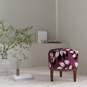 Può includere: Un pouf rotondo con un tessuto bordeaux scuro con un motivo a foglie bianche. Il pouf ha una base e gambe in legno. Un vaso con rami verdi e libri sono sullo sfondo. Il diametro del pouf è di circa 40 cm.