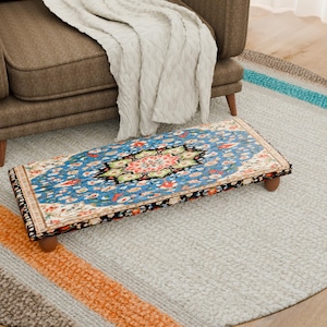 Puede incluir: Un reposapiés rectangular con un estampado floral azul, rojo y verde se encuentra sobre una alfombra gris claro. El reposapiés tiene patas de madera y un borde negro. Un sillón marrón y una manta blanca están en el fondo.