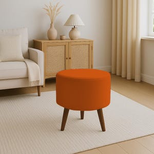 May include: An orange, round ottoman with three dark brown wooden legs. The ottoman is in a living room setting, with a cream-colored rug and a light-colored cabinet in the background.