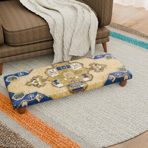 Puede incluir: Un reposapiés rectangular con un diseño estampado en tonos beige, azul y dorado. El reposapiés tiene patas de madera y está sobre una alfombra gris claro. Un sillón marrón y una manta crema están en el fondo. Mide unos 76 cm de largo.