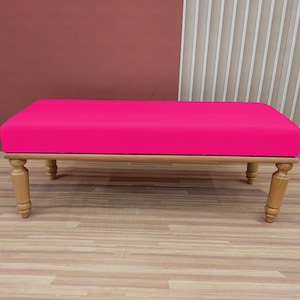 Puede incluir: Un banco tapizado de color rosa con patas de madera. El banco está sobre un suelo de madera.
