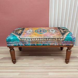 May include: A wooden ottoman with a colorful patterned fabric top. The fabric features a geometric design in shades of teal, pink, yellow, and brown.