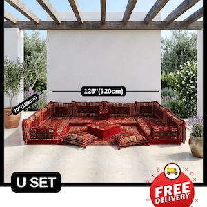 Op de afbeelding: Een rode U-vormige loungeset voor buiten, 320 cm breed en 180 cm diep, met kussens met patronen en een centrale vierkante poef. De set staat op een betegeld terras, met een witte muur en houten balken erboven.