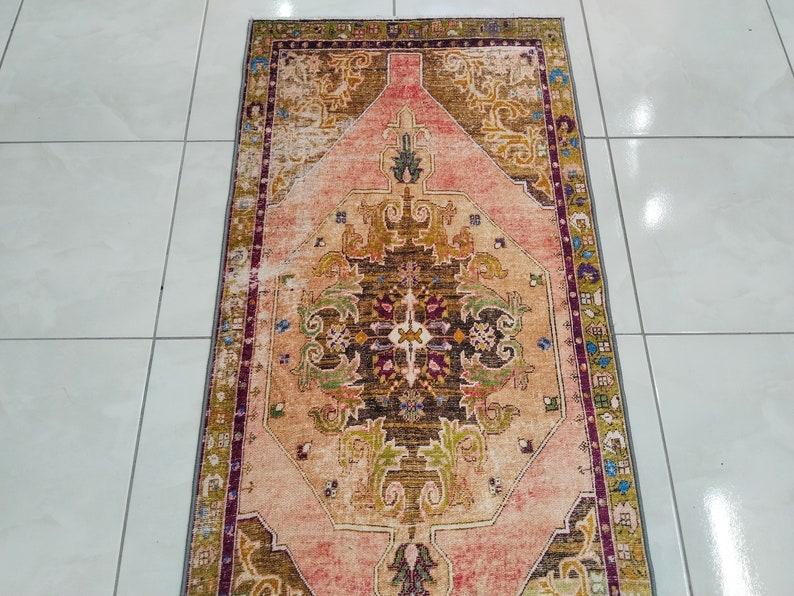 K&ouml;nnte beinhalten: Ein Vintage-Teppich mit einem floralen Muster in Rosa, Braun und Gr&uuml;n. Der Teppich hat einen verblassten, abgenutzten Look und ist perfekt, um Ihrem Zuhause einen Hauch von Boho-Stil zu verleihen.