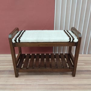 May include: A wooden bench with a cream-colored cushion and dark brown trim. The bench has a slatted shelf below the seat and is made of dark brown wood. The cushion is decorated with a dark brown zigzag pattern.