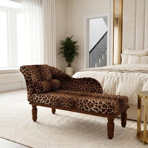May include: A chaise lounge upholstered in a brown and black leopard print fabric. The furniture piece features a curved backrest, a matching cylindrical bolster pillow, and four dark brown wooden legs with a turned, spindle-style design.