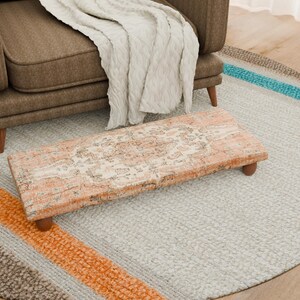 Puede incluir: Un reposapiés rectangular con una parte superior estampada en melocotón y crema desgastado y patas de madera oscura. Se encuentra sobre una alfombra redonda gris claro con rayas naranjas, azules y marrones. Un sillón marrón y una manta crema están en el fondo.