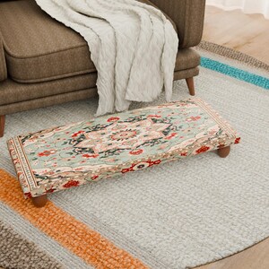 Peut inclure: Un pouf rectangulaire avec un dessus en tissu à motifs dans les tons de turquoise, de rouge et de crème, reposant sur quatre pieds en bois. Le pouf est posé sur un tapis gris clair avec des rayures colorées.