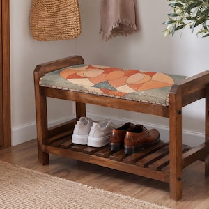 May include: A wooden shoe bench with a rectangular frame and a slatted lower shelf. The top features a cushioned seat upholstered in a fabric with a geometric pattern in shades of orange, tan, and muted green. Two pairs of shoes sit on the shelf.