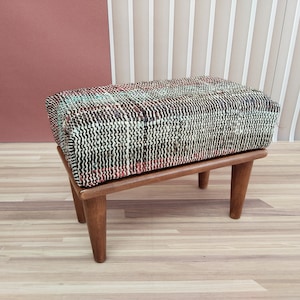 Puede incluir: Un pequeño taburete de madera con un asiento de tejido tejido en tonos marrones, negros, blancos y rojos. El taburete tiene tres patas y está sentado en un suelo de madera.