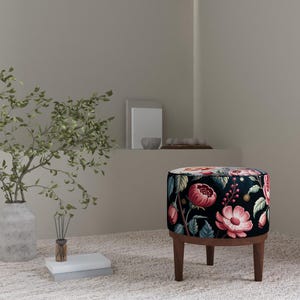 May include: A round ottoman with a floral pattern in pink, red, and green on a black background. The ottoman has dark wooden legs and is placed on a light-colored rug. A vase with greenery and other decorative items are in the background.
