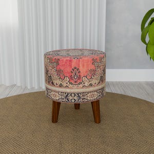 Op de afbeelding: Een ronde, roze en blauwe gepatroneerde poef met houten poten. De poef staat op een bruin vloerkleed.
