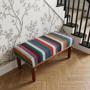 a colorful bench with a striped cushion, placed in front of a wall with a floral pattern. The bench has wooden legs and is situated on a wooden floor.