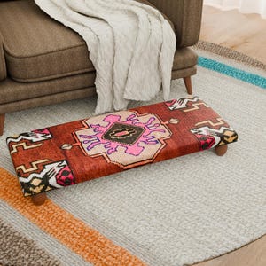 Puede incluir: Un otomán rectangular con un diseño de alfombra tradicional en tonos rojos, rosas y beige. El otomán tiene patas cortas de madera redondeadas y está colocado sobre una alfombra gris claro. Un sillón marrón y una manta blanca están en el fondo.