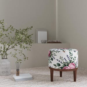 May include: A round ottoman with a floral pattern featuring pink roses, purple lavender, and green leaves on a white background. The ottoman has a dark brown wooden base and legs. The room has a neutral color palette.