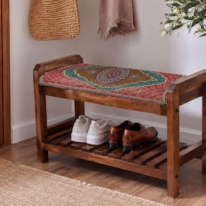 Può includere: Una panca scarpiera in legno con telaio rettangolare e ripiano inferiore a doghe. La seduta superiore presenta un cuscino in tessuto colorato con un dettagliato motivo ispirato ai mandala nei toni del rosso, verde e oro. Un paio di scarpe da ginnastica bianche e scarpe in pelle marrone poggiano sul ripiano.