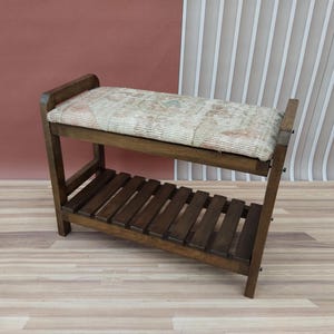 Puede incluir: Un banco de madera con un asiento acolchado y un estante inferior. El cojín está cubierto con una tela estampada en tonos beige, rosa y marrón. El banco está hecho de madera marrón oscuro y tiene un diseño simple y rústico.