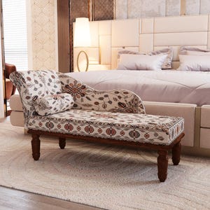 May include: A chaise lounge with a floral pattern in a bedroom setting. The chaise has a wooden frame and legs, and a patterned cushion with a matching bolster pillow. The room has a neutral color palette with a large bed in the background.