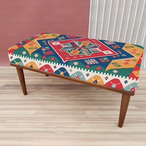Op de afbeelding: Een kleurrijke, gepatroneerde ottomaan met een houten frame. De ottomaan heeft een geometrisch ontwerp met de kleuren rood, blauw, geel, groen en wit.