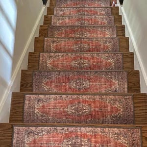 May include: A set of rectangular stair treads featuring a faded red and beige medallion pattern. Each tread is centered on a dark brown wooden stair step, creating a repeating visual sequence that leads down the length of the staircase.