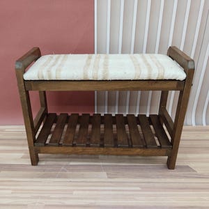 May include: A wooden bench with a cushioned top and a lower shelf. The bench is brown and the cushion is white with brown stripes.