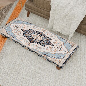 Puede incluir: Un otomán rectangular con un diseño de alfombra detallado y ornamentado. La alfombra presenta un medallón central en azul marino, crema y naranja, enmarcado por un borde azul claro. El otomán descansa sobre cuatro patas cortas de madera.