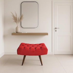 Peut inclure: Un pouf rouge capitonné avec des pieds en bois foncé. Il est placé dans une pièce avec un miroir, une étagère et une porte blanche. Un élément d'accent vif.