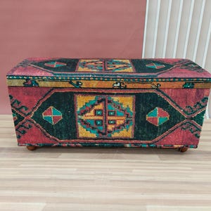 Può includere: Un pouf di stoccaggio rettangolare con una copertura in tessuto a motivi colorati. Il tessuto presenta un design di tappeto tradizionale con colori rosso, verde, blu e giallo.