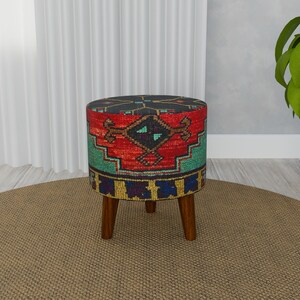Può includere: Un pouf colorato e con motivi con un designo rosso, blu e verde. Il pouf ha quattro gambe in legno e si trova su un tappeto marrone.
