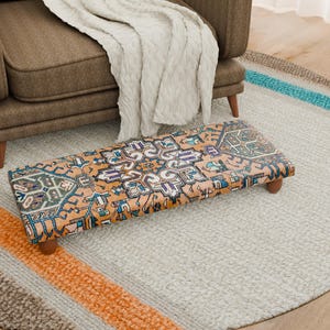 Puede incluir: Un reposapiés rectangular con un diseño estampado en tonos naranja, azul y morado. El reposapiés tiene patas de madera y está colocado sobre una alfombra gris claro con un borde a rayas de colores. Un sillón marrón y una manta crema están en el fondo. Mide unos 90 cm de largo.