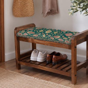 May include: A wooden shoe bench with a dark brown finish and a rectangular seat cushion featuring a dark green fabric with a multi-colored floral pattern. The bench has a slatted lower shelf holding a pair of white sneakers and brown leather shoes.