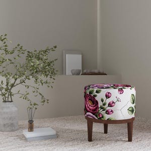 May include: A small, round ottoman with a floral pattern featuring large, deep pink roses and green leaves on a white background. The ottoman has dark brown wooden legs and a matching wooden base.