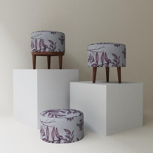 May include: Three small, round ottomans with wooden legs. The ottomans have a light gray fabric with a purple floral pattern. Two ottomans are elevated on white pedestals. The smallest ottoman sits on the floor.