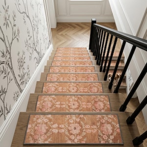 May include: A set of rectangular stair treads featuring a burnt orange background with a repeating light pink floral pattern. Each tread has a dark gray border and is installed on wooden stairs next to a black railing and floral wallpaper.
