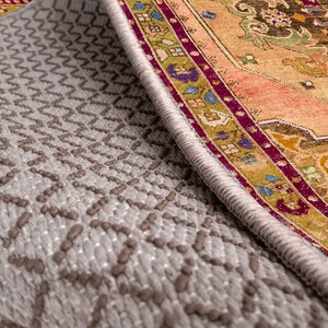 K&ouml;nnte beinhalten: Nahaufnahme der Unterseite eines Teppichs mit einer grauen, strukturierten R&uuml;ckseite und einem bunten, gemusterten Rand. Der Rand zeigt ein sich wiederholendes Muster aus floralen Motiven und geometrischen Formen in Rot-, Orange-, Gelb-, Gr&uuml;n-, Blau- und Violettt&ouml;nen.