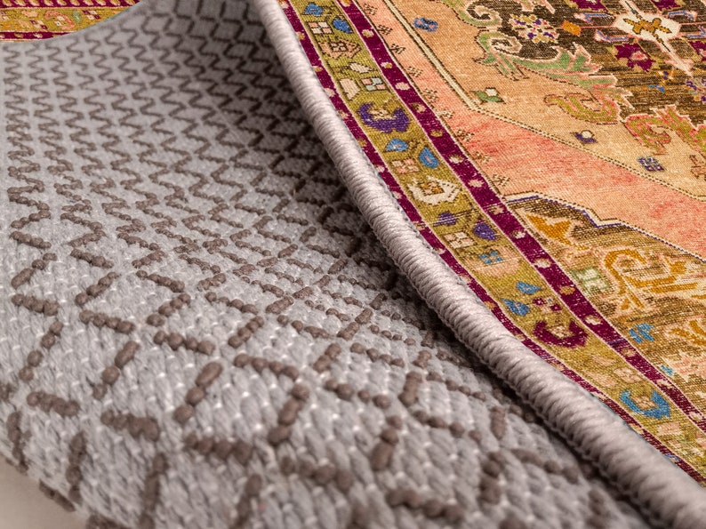 K&ouml;nnte beinhalten: Nahaufnahme der Unterseite eines Teppichs mit einer grauen, strukturierten R&uuml;ckseite und einem bunten, gemusterten Rand. Der Rand zeigt ein sich wiederholendes Muster aus floralen Motiven und geometrischen Formen in Rot-, Orange-, Gelb-, Gr&uuml;n-, Blau- und Violettt&ouml;nen.