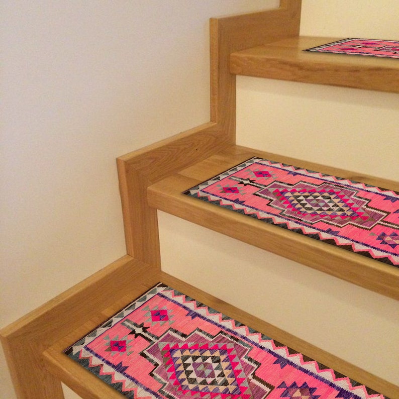 May include: Pink and patterned stair treads with a geometric design are placed on wooden stairs. The treads feature a central diamond pattern in shades of pink, purple, and black, with a border of triangles and zigzags.
